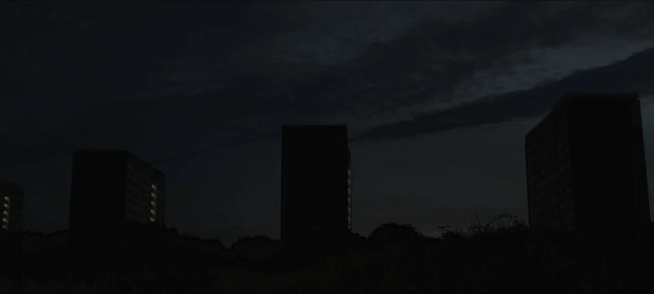 Containment (2015) movie still: wide — A dark urban landscape shows several tall residential blocks against a black sky. These apartment structures…