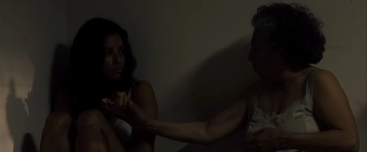 Frontera (2014) movie still: medium — Two women sit together in the corner of a dimly lit room against white walls. The older woman holds the hand…