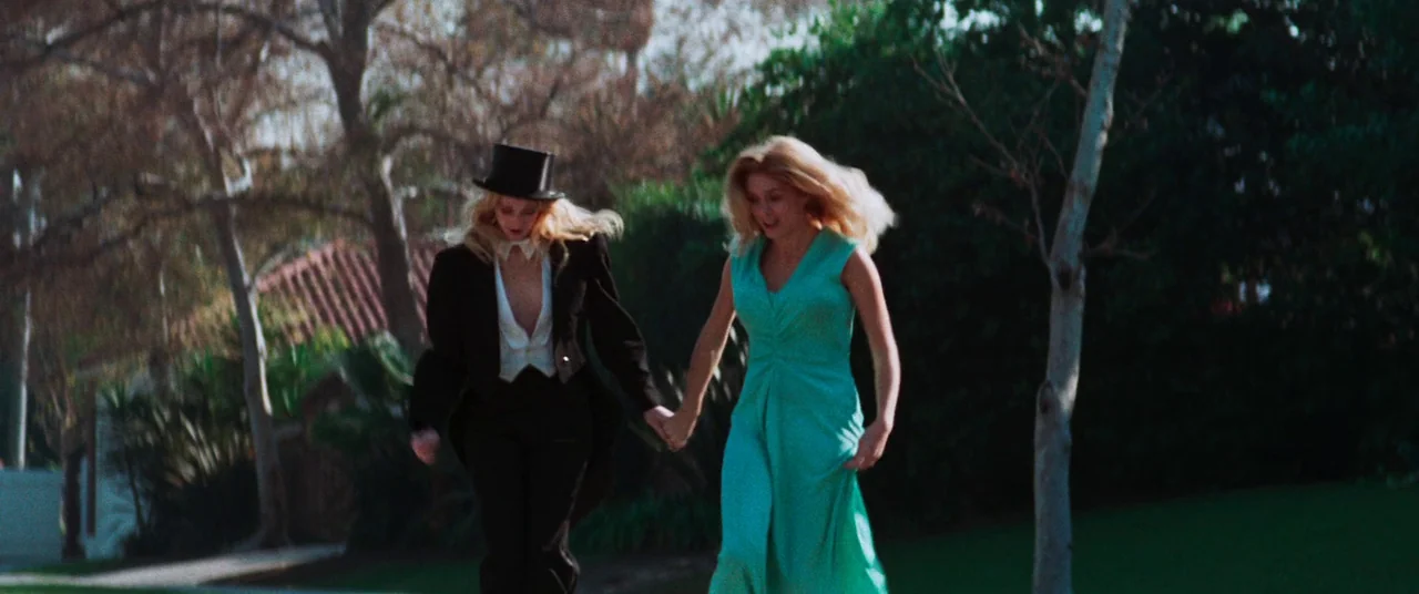 Death Game (1977) movie still: fullbody — Two women run along a tree-lined road during the daytime. One woman wears a tuxedo and top hat, while the…
