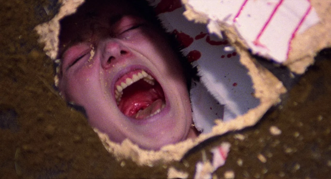 City of the Living Dead (1980) movie still: closeup — A person's face is framed within a jagged hole broken through a drywall partition. The person…