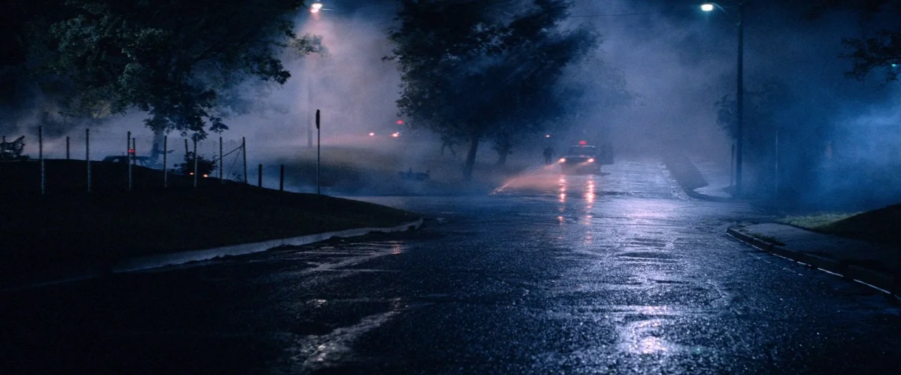 The Outsiders (1983) movie still: wide — A dark residential suburban street is covered in thick fog at night. A lone police vehicle slowly…