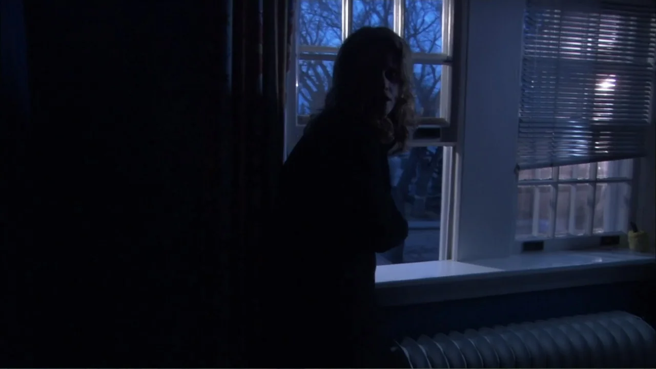 O código oculto (2025) movie still: medium — A dark room features a window with white blinds overlooking a night scene. A woman stands by the window…