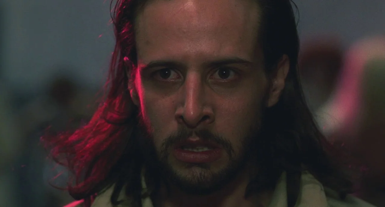 Santa Sangre (1989) movie still: closeup — A dimly lit space features a man captured in a tight portrait composition. The subject stares forward…