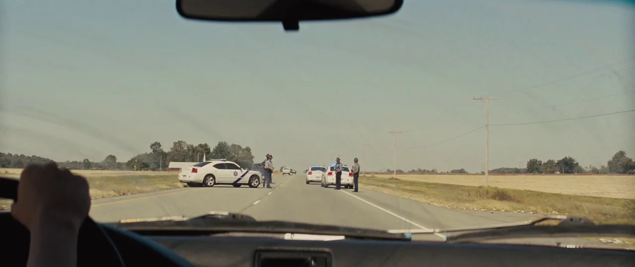 Mud (2012) movie still: wide — A flat stretch of rural highway under a pale, cloudless sky. Three uniformed state troopers stand beside two white…