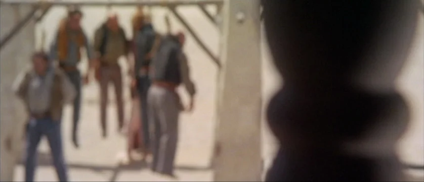 Django Kill (1967) movie still: wide — A dusty outdoor area contains a wooden gallows structure in the foreground. Several men stand in a group,…