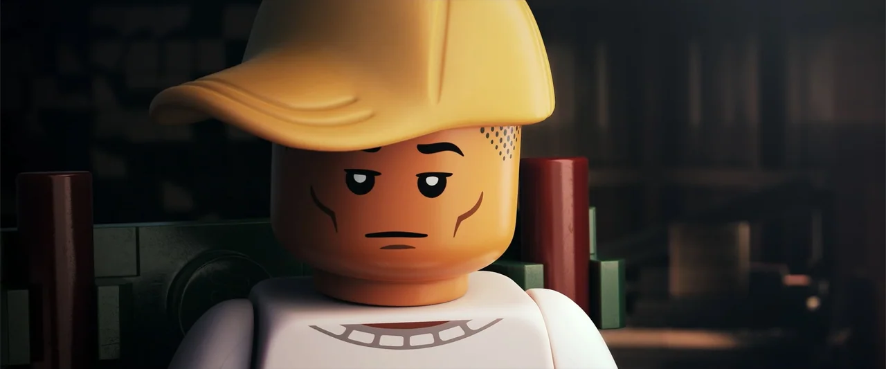Piece by Piece (2024) movie still: closeup — A close-up shot of a plastic minifigure character in a workspace environment. The figure wears a yellow…