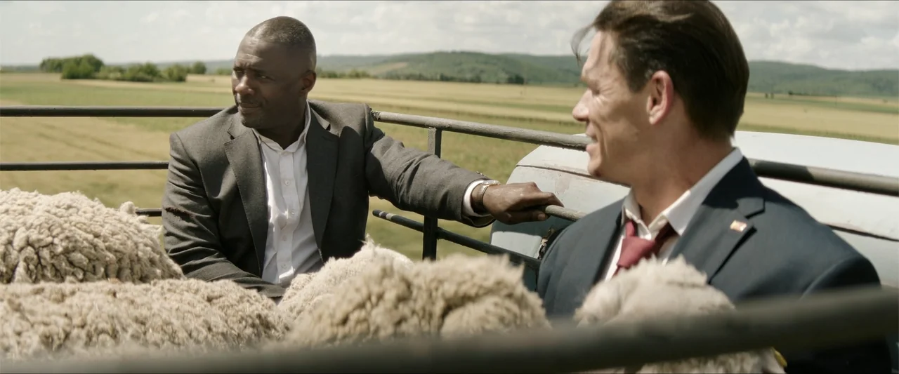 Heads of State (2025) movie still: medium — Two men in business suits ride in the open cargo bed of a truck. The vehicle moves past rolling fields…
