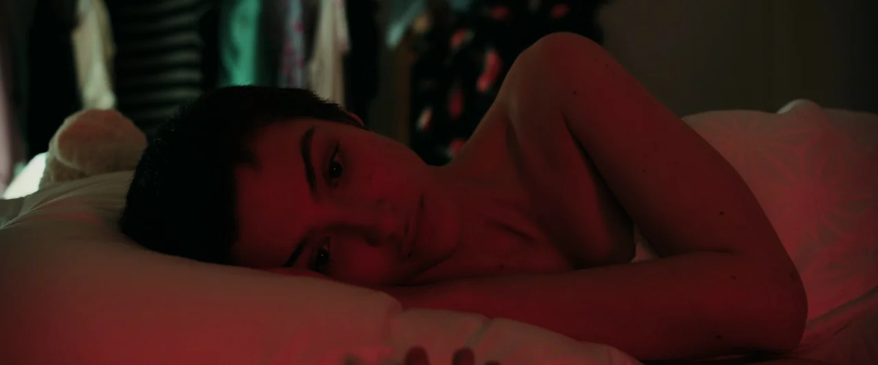 Anna (2019) movie still: closeup — A person lies on a white pillow in a dark space with hanging clothes visible. The subject is partially covered by…