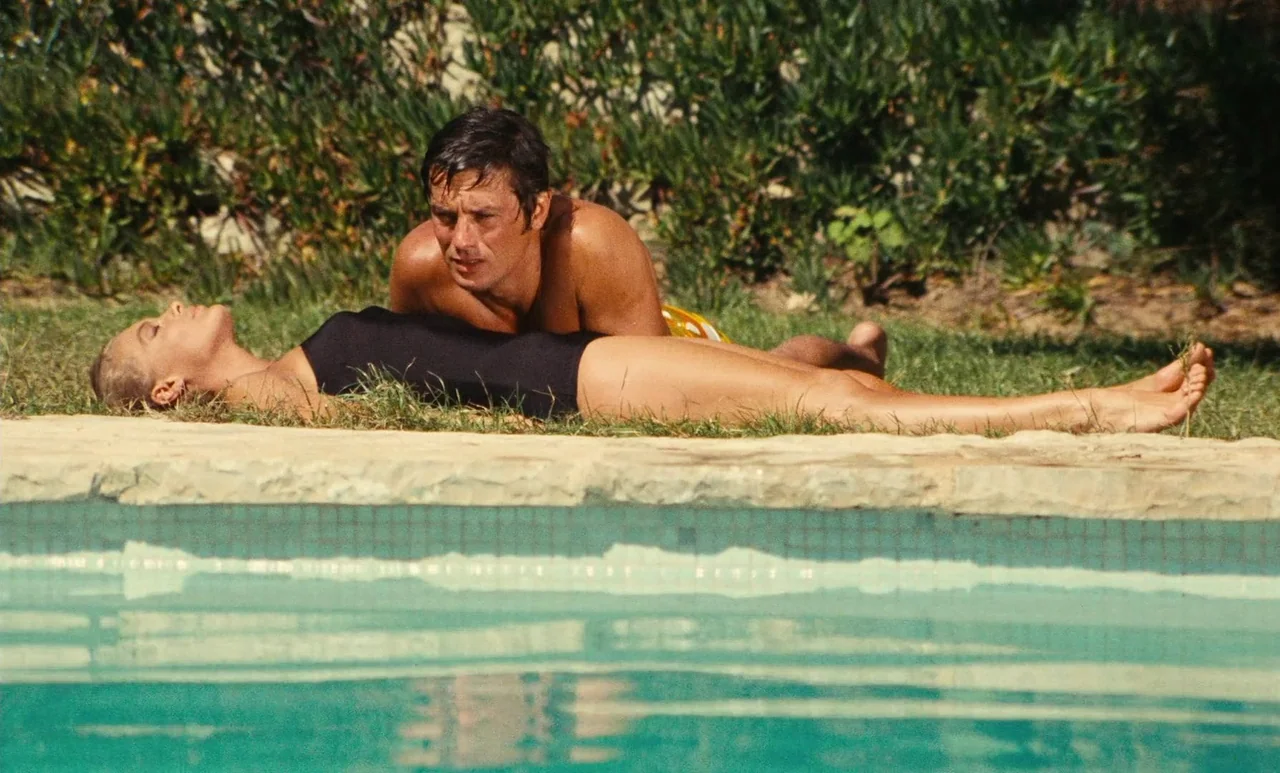 The Swimming Pool (1969) movie still: medium — Two people lie on the grass beside a stone-edged pool under the sun. The man is behind the woman,…