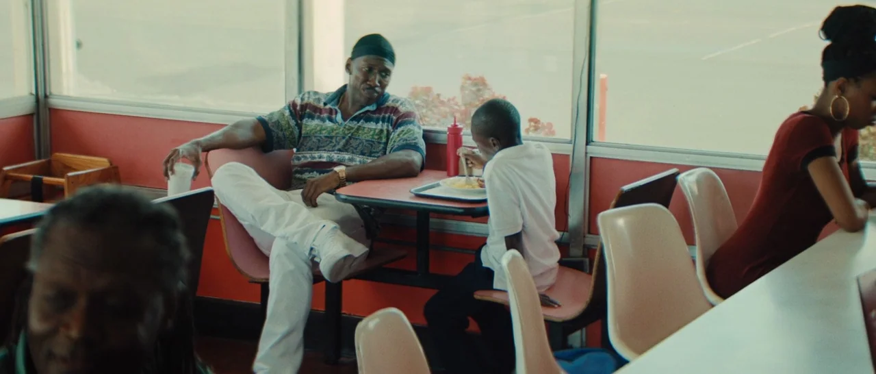 Moonlight (2016) movie still: medium — A modest diner booth features a red-padded bench and a rectangular Formica table. An adult man and a young…