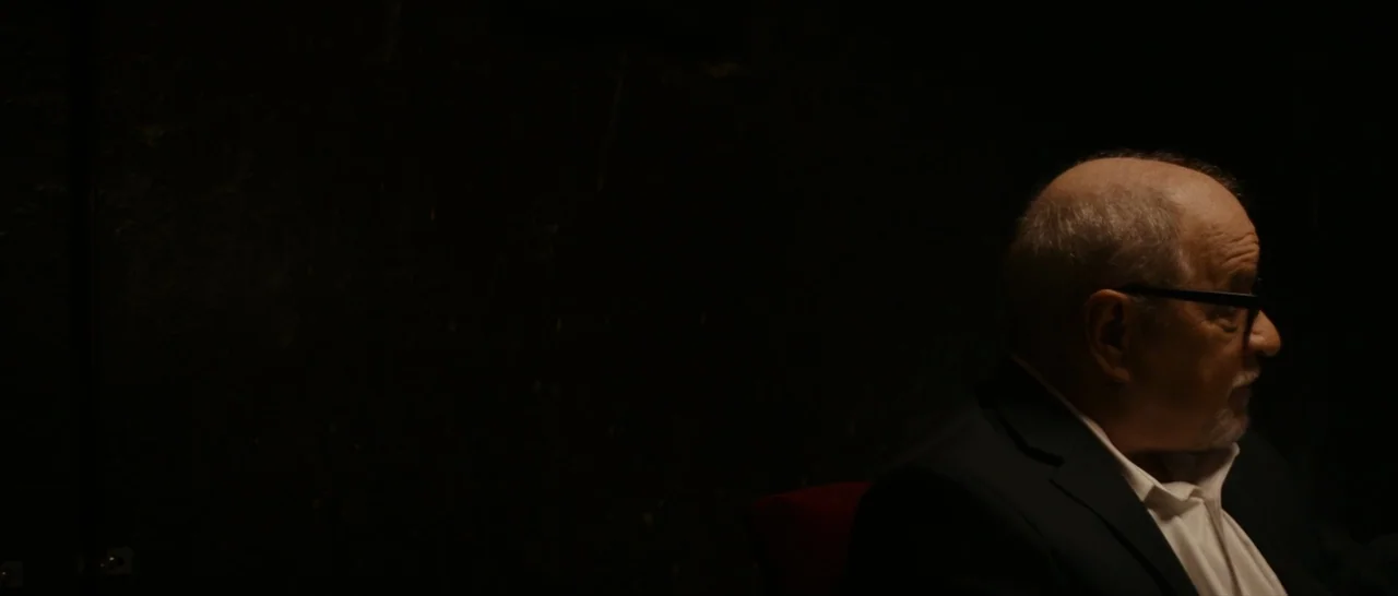 Dog Eat Dog (2016) movie still: medium — A dark, minimally lit interior space with a black textured wall background. A man in a suit is seated and…