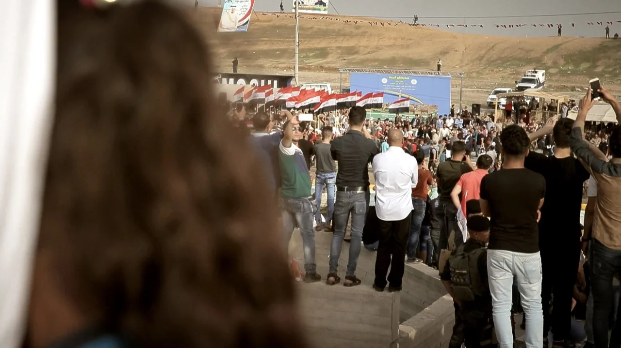City of Two Springs (2019) movie still: medium — An outdoor dusty plaza with a crowd facing a distant stage with banners. Men in casual clothes…
