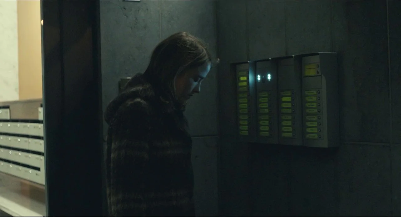 The Unknown Girl (2016) movie still: medium — A dimly lit modern apartment lobby features dark stone walls and a metal intercom panel. A young woman…