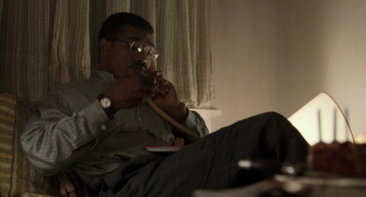 One False Move (1991) movie still: medium — A man sits on a bed in a dim motel room with patterned curtains behind him. He holds a telephone to his…