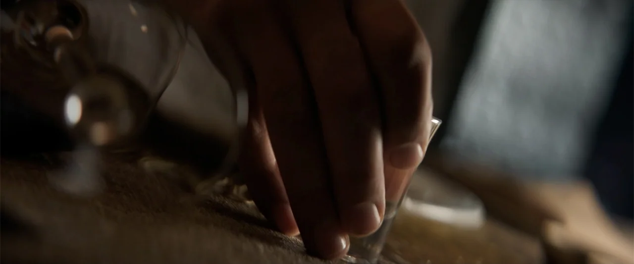 Drop (2025) movie still: closeup — A dark wooden surface holds a small glass under low directional illumination. A human hand rests firmly against…
