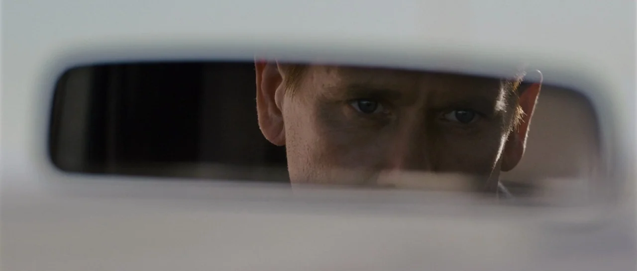 Cop Car (2015) movie still: closeup — The interior of a vehicle seen through the perspective of a rear view mirror. A man gazes directly into the…