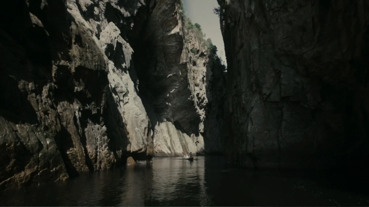 The Dinosaur Project (2012) movie still: establishing — A narrow waterway flows between two high, jagged rock canyon walls. A lone boat sits in the…