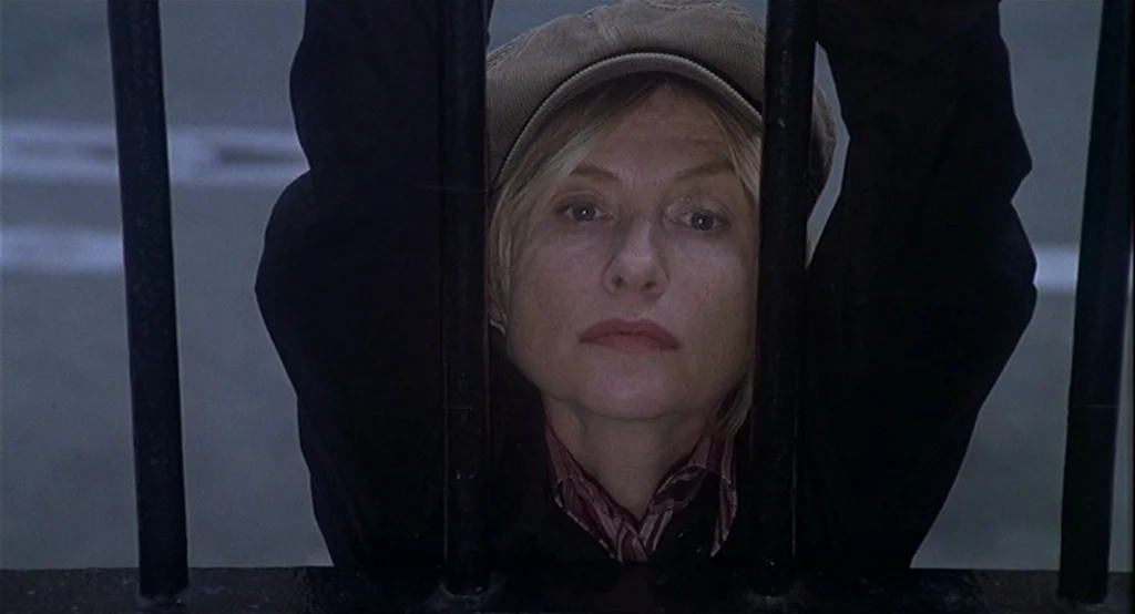 Deux (2002) movie still: closeup — A woman looks through a series of vertical black iron bars in an outdoor setting. Her hands are visible as she…