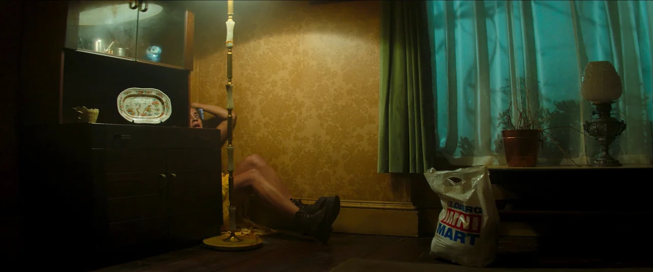 Unwelcome (2022) movie still: medium — A dimly lit room features patterned yellow wallpaper and an antique wooden cabinet unit. A woman sits on the…