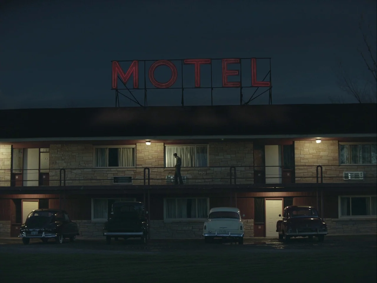 The Mountain (2018) movie still: wide — A two-story motor lodge at night features stone masonry and vintage automobiles parked underneath. A lone…