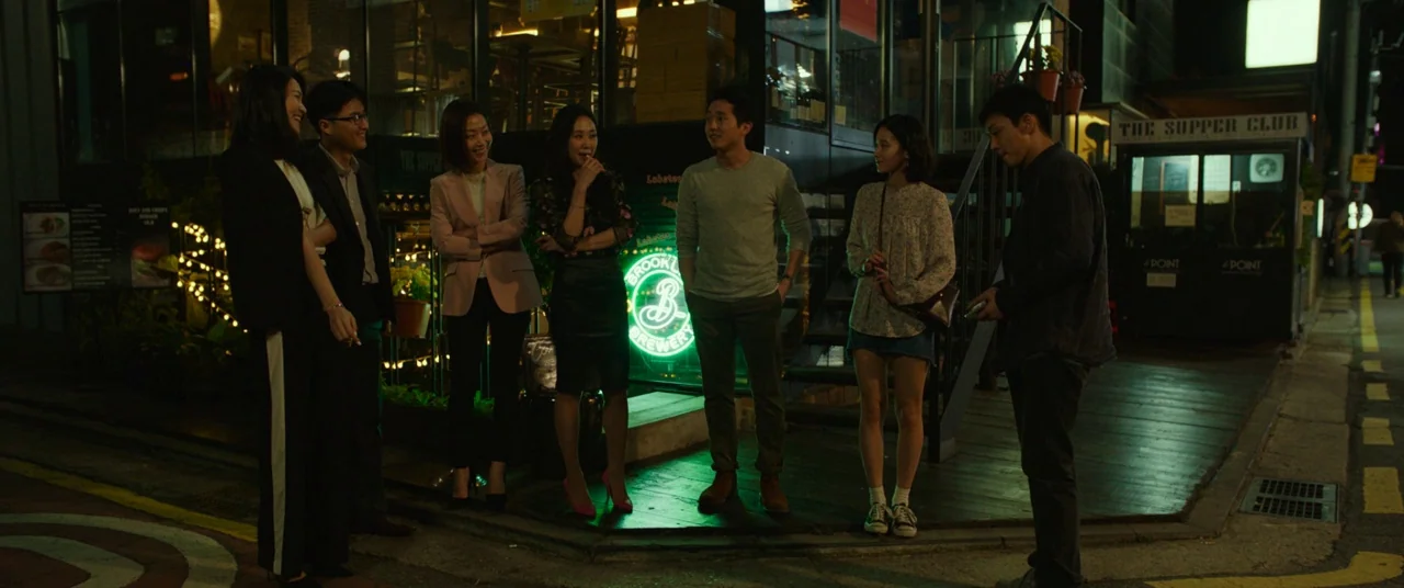 Burning (2018) movie still: fullbody — A group of friends stands on a dark city street outside a glass-fronted establishment. The group interacts…