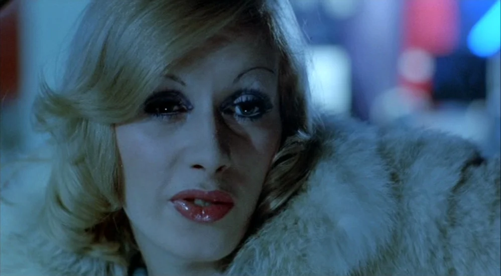 Un Flic (1972) movie still: closeup — A dimly lit interior space with blurred neon lights visible in the background. A woman in a white fur coat…