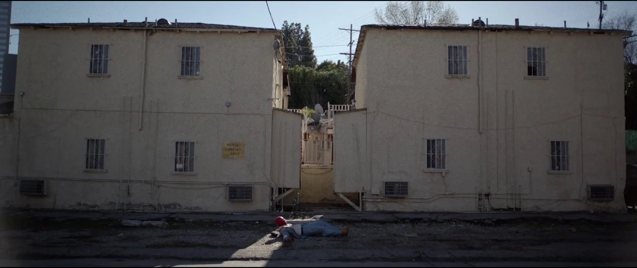 Lowlife (2017) movie still: wide — A split, two-story beige stucco apartment complex features a narrow dark alleyway between buildings. A single…