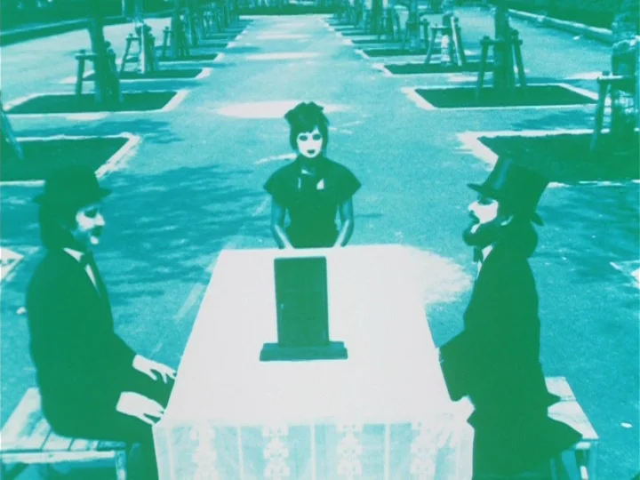 Labyrinth Tale (1975) movie still: wide — A long paved pathway lined with young trees enclosed in wooden stakes. Two men in formal suits and a woman…