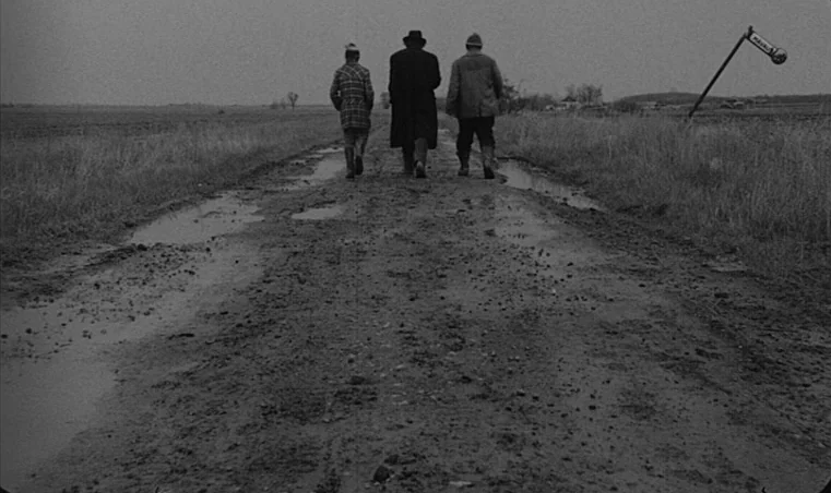 Sátántangó (1994) movie still: wide — A desolate dirt road cuts through flat, grassy fields under a grey, overcast sky. Three men walk away from the…