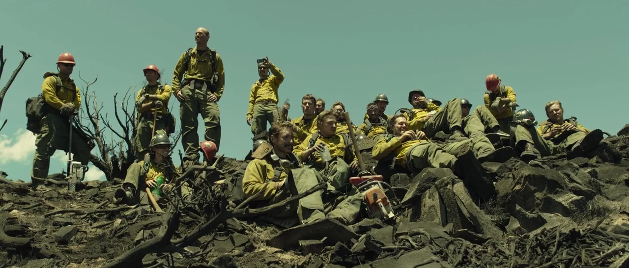 Only The Brave (2017) movie still: wide — A group of wildland firefighters rests on a desolate, rock-covered hillside after a blaze. The ground is…
