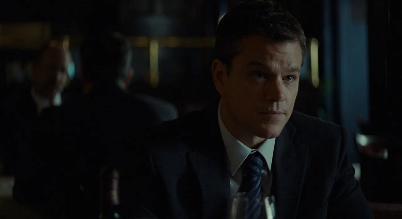 Promised Land (2012) movie still: medium — A dimly lit restaurant interior with dark surfaces and blurred background patrons. A man in a business…