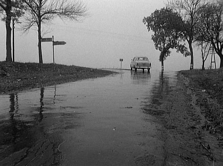 Knife In The Water (1962) movie still: wide — A wet asphalt road leads toward a horizon under an overcast grey sky. A lone sedan travels away into…