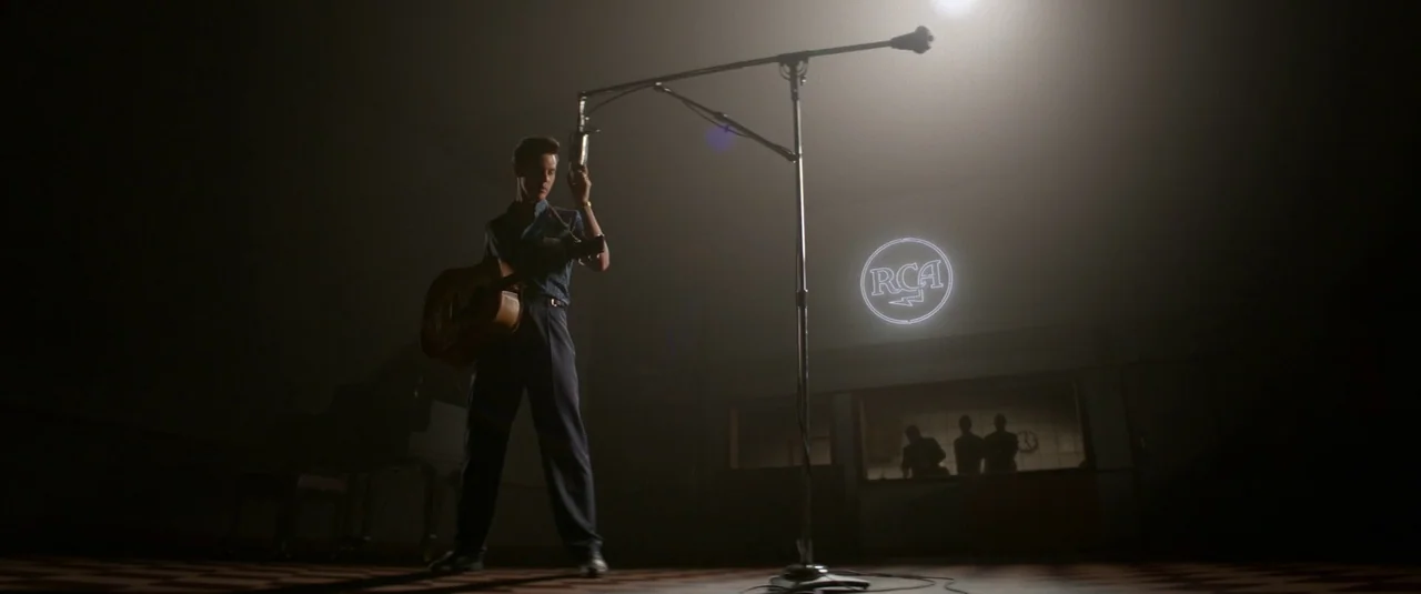 Elvis (2022) movie still: fullbody — A dimly lit recording studio features a man standing in front of a microphone. Through a window in the…
