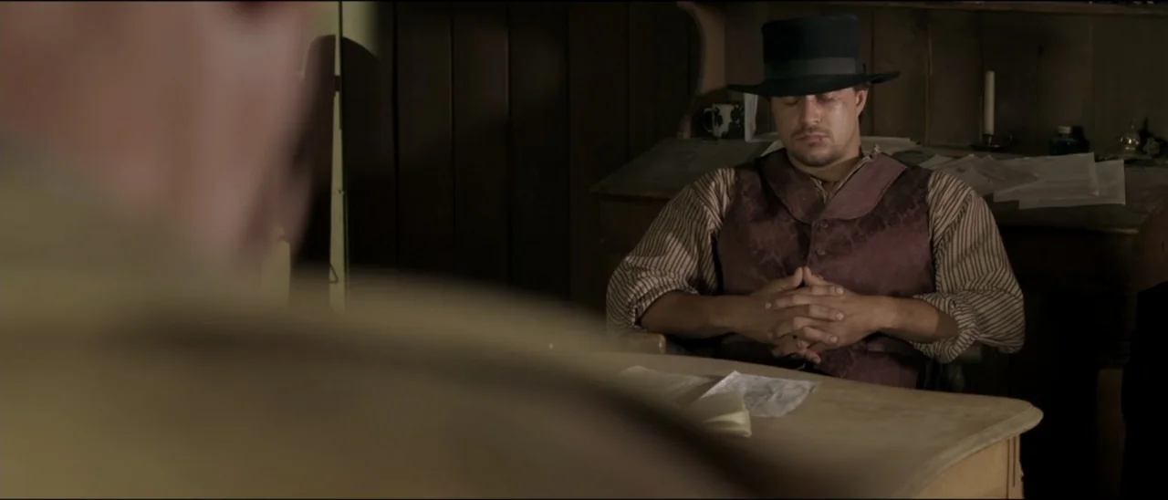 Dead for a Dollar (2022) movie still: medium — A dimly lit room features wood-paneled walls and a desk covered with scattered papers. A man wearing…