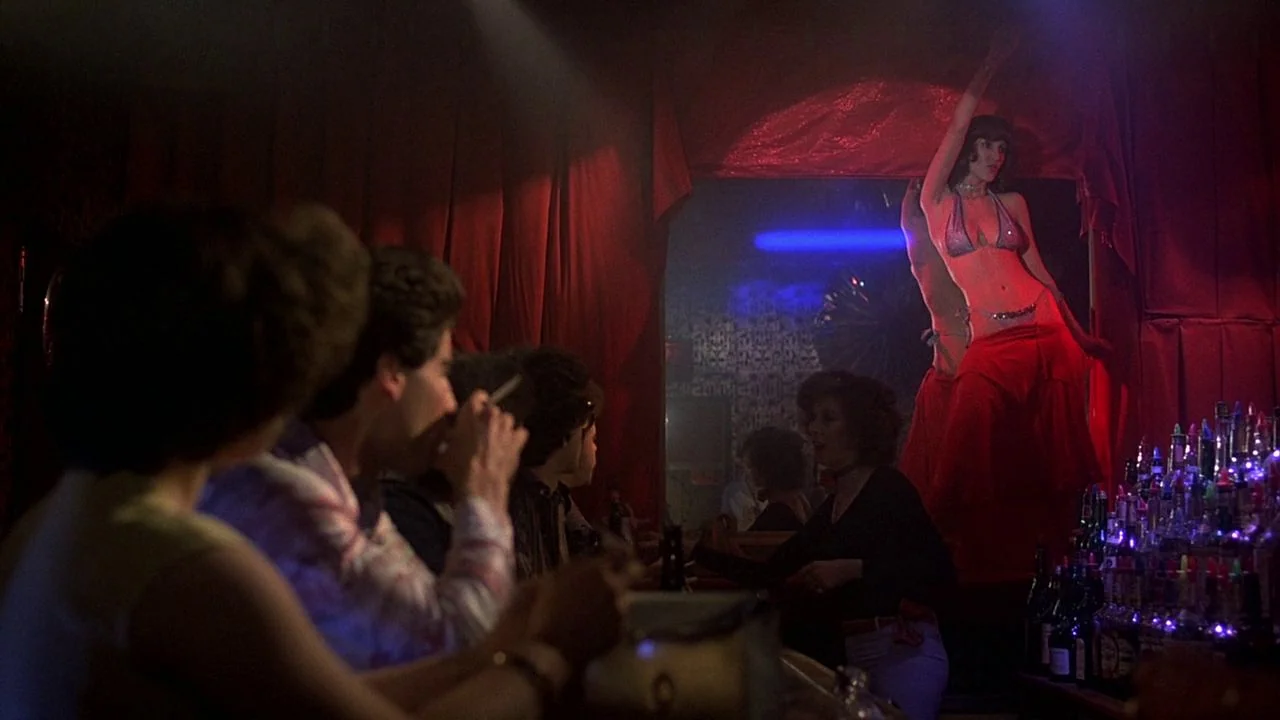 Saturday Night Fever (1977) movie still: medium — A dimly lit club with a red-curtained stage and a crowded bar area. A dancer performs on stage…
