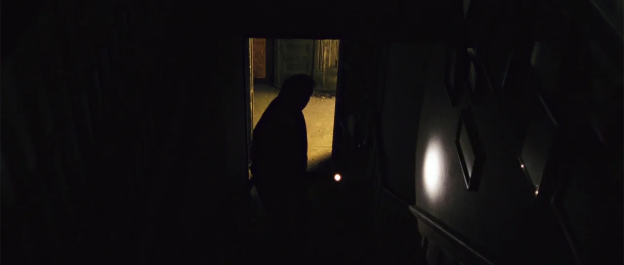 The New Daughter (2009) movie still: medium — A dark stairwell with a silhouette standing in a doorway at the top landing. The person holds a small…