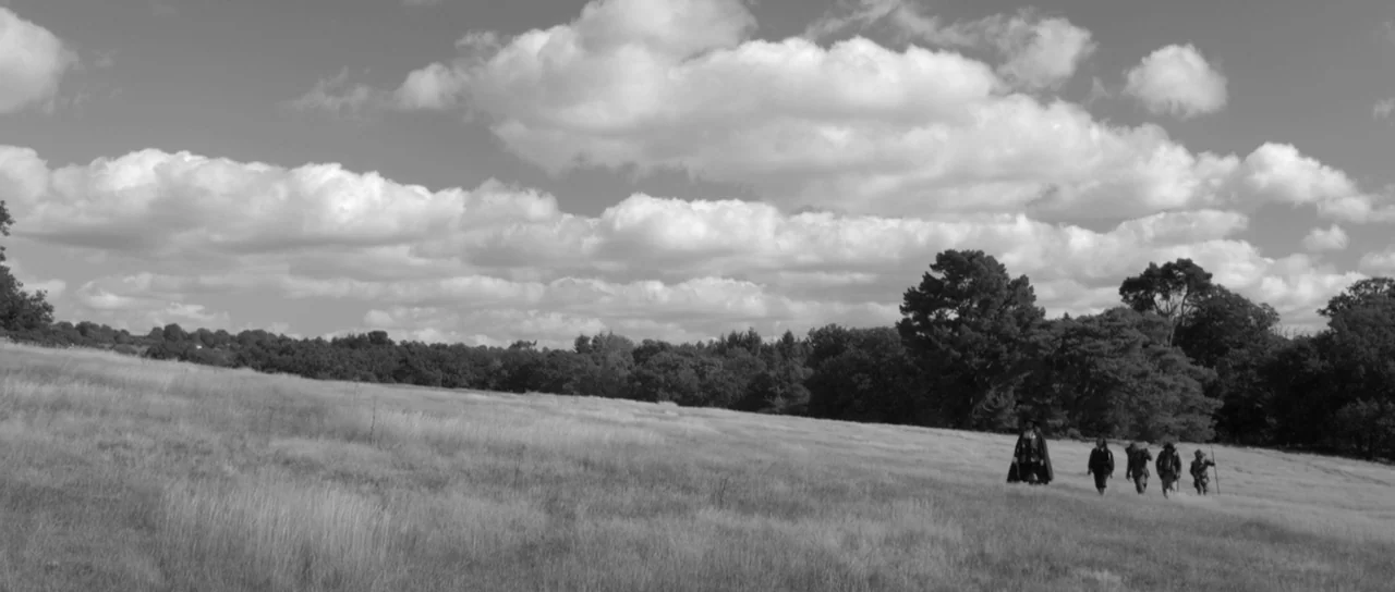 A Field In England (2013) movie still: wide — An expansive meadow features dry grasses under a sky filled with bright white clouds. A small group of…