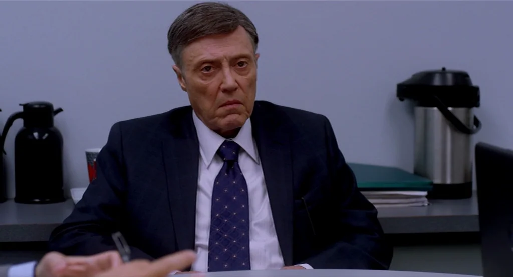 Dark Horse (2011) movie still: medium — A man in a suit sits behind a grey conference table in a bland office. Two large thermal beverage carafes…