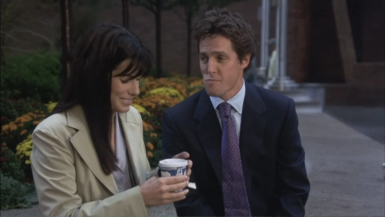 Two Weeks Notice (2002) movie still: medium — A paved outdoor plaza with manicured flower beds and trees in the background. A woman sits next to a…