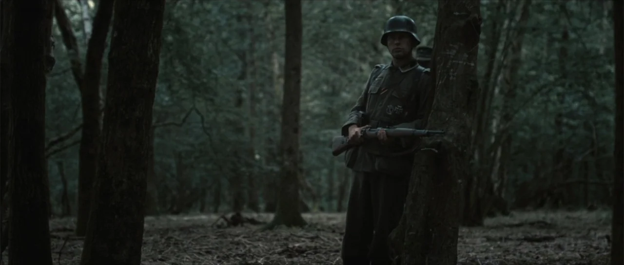 Sin retorno (2009) movie still: fullbody — A dense woodland environment with tall trees and thick foliage under an overcast sky. A soldier in…