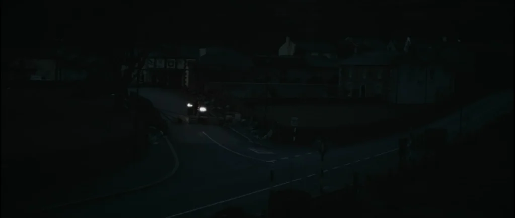 One Hundred Mornings (2009) movie still: wide — A dark rural intersection at night with stone houses visible in the background. A vehicle sits at…