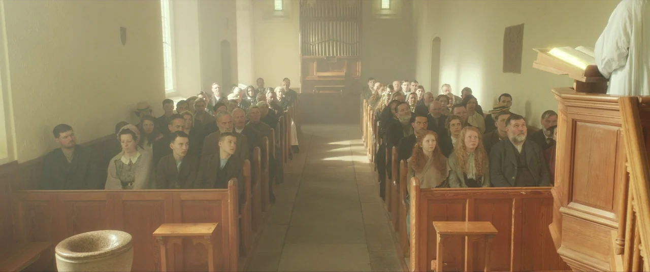 Sunset Song (2015) movie still: wide — A congregation sits in wooden pews inside a light-filled, sparse church interior. The members face the front…
