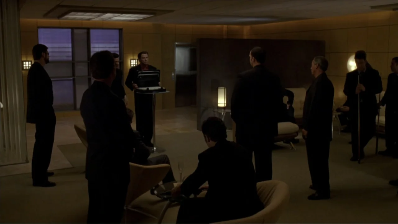 The Punisher (2004) movie still: wide — A spacious modern lounge area features tan leather furniture and dark wall panels. Several men in formal…