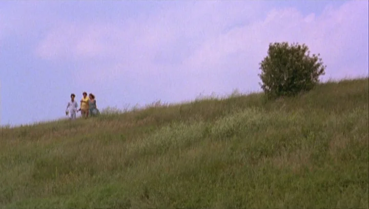 Highway 61 (1991) movie still: wide — A grassy hill extends toward a blue sky with a single small bush present. Three people walk across the incline…