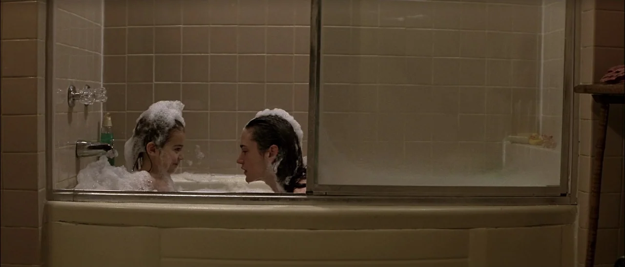 Dark Water (2005) movie still: medium — A bathroom with beige tiled walls features a glass shower door enclosing a bathtub. A young woman and a…