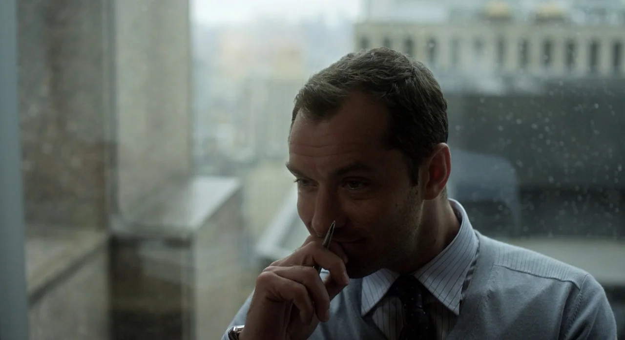 Side Effects (2013) movie still: closeup — An interior space with a window overlooking a blurred cityscape. A man in a sweater stands looking toward…