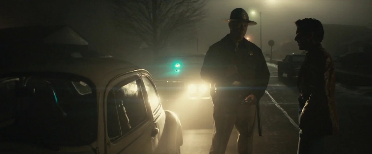 Extremely Wicked, Shockingly Evil and Vile (2019) movie still: fullbody — A foggy night on a dark road with a patrol car parked nearby. A uniformed…