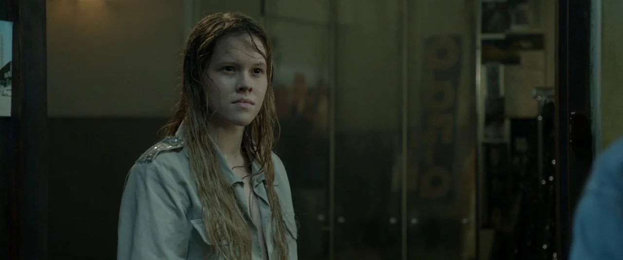 The Lure (2015) movie still: medium — A dimly lit indoor space features a dark corridor with reflective door panels. A young woman with wet,…