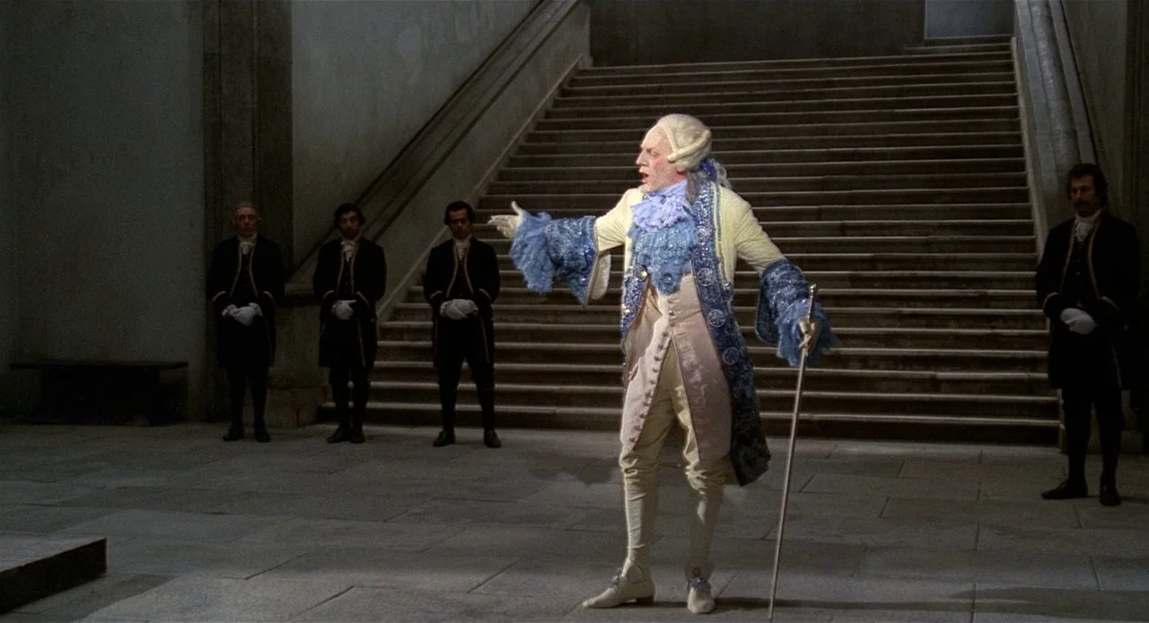 Fellini’s Casanova (1976) movie still: fullbody — An ornate palace interior features a wide central stone staircase and stone floor. A central…
