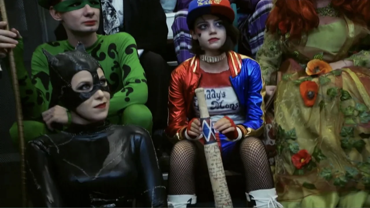 Reign of the Superwomen (2021) movie still: medium — A group of people dressed as various comic book characters sit inside a train car. They remain…